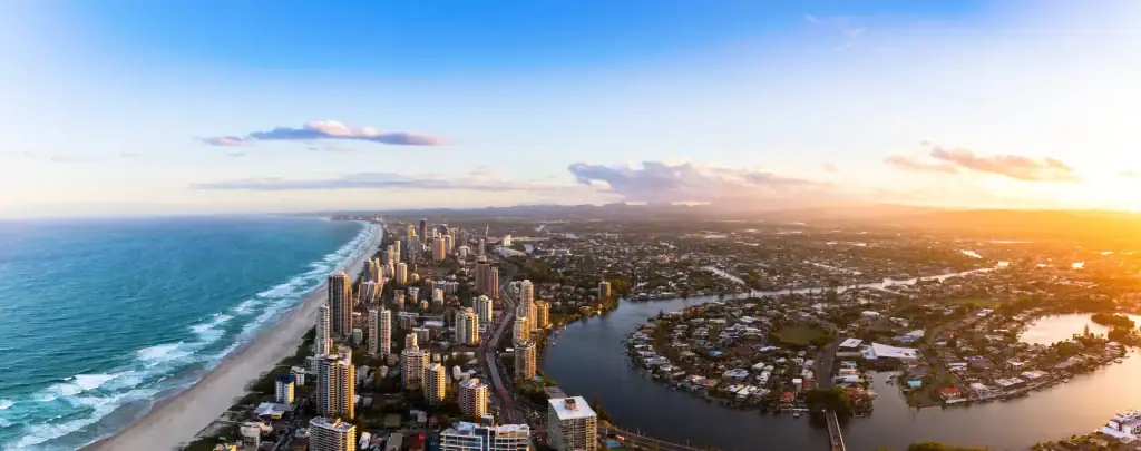 Aerial sunset panorama of Gold Coast Surfers Paradise skyscrapers lining a long sandy beach, with canal waterways and suburban sprawl stretching inland